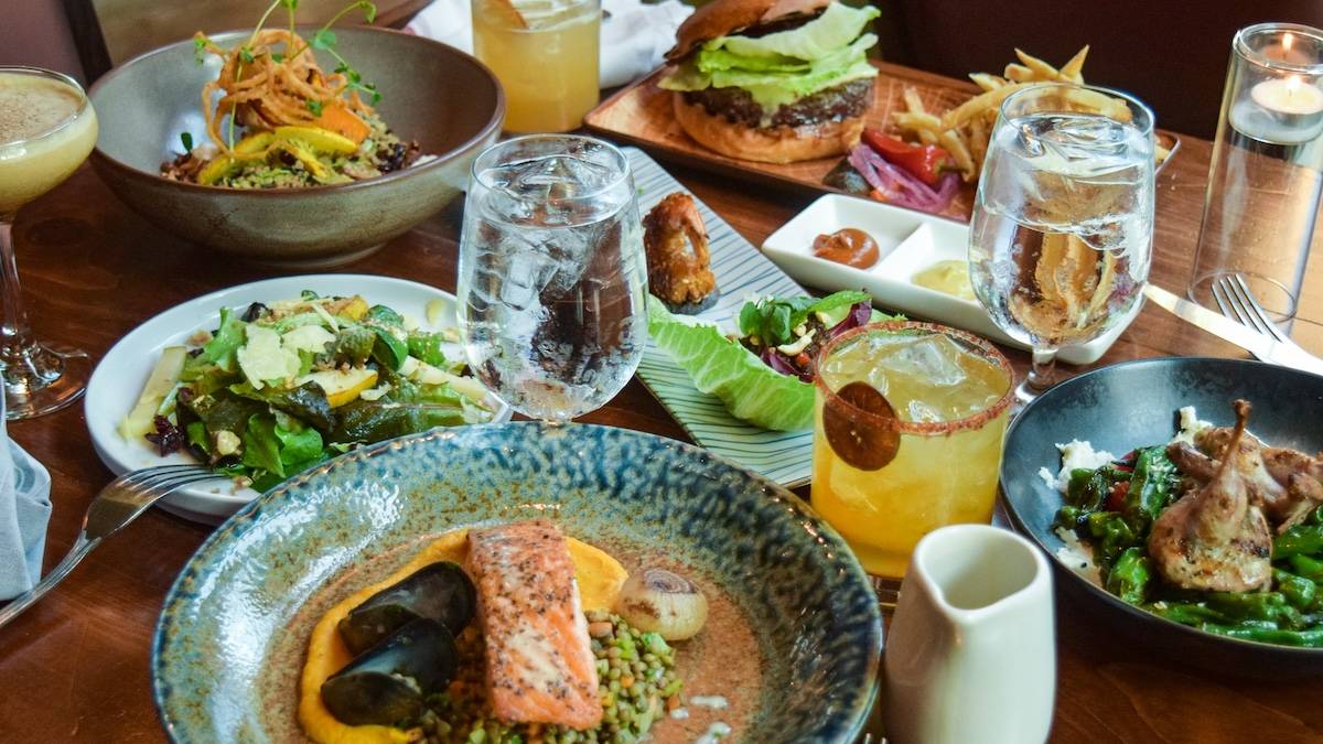 Several plates with a variety of food on a wooden table surrounded by drinks and cutlery
